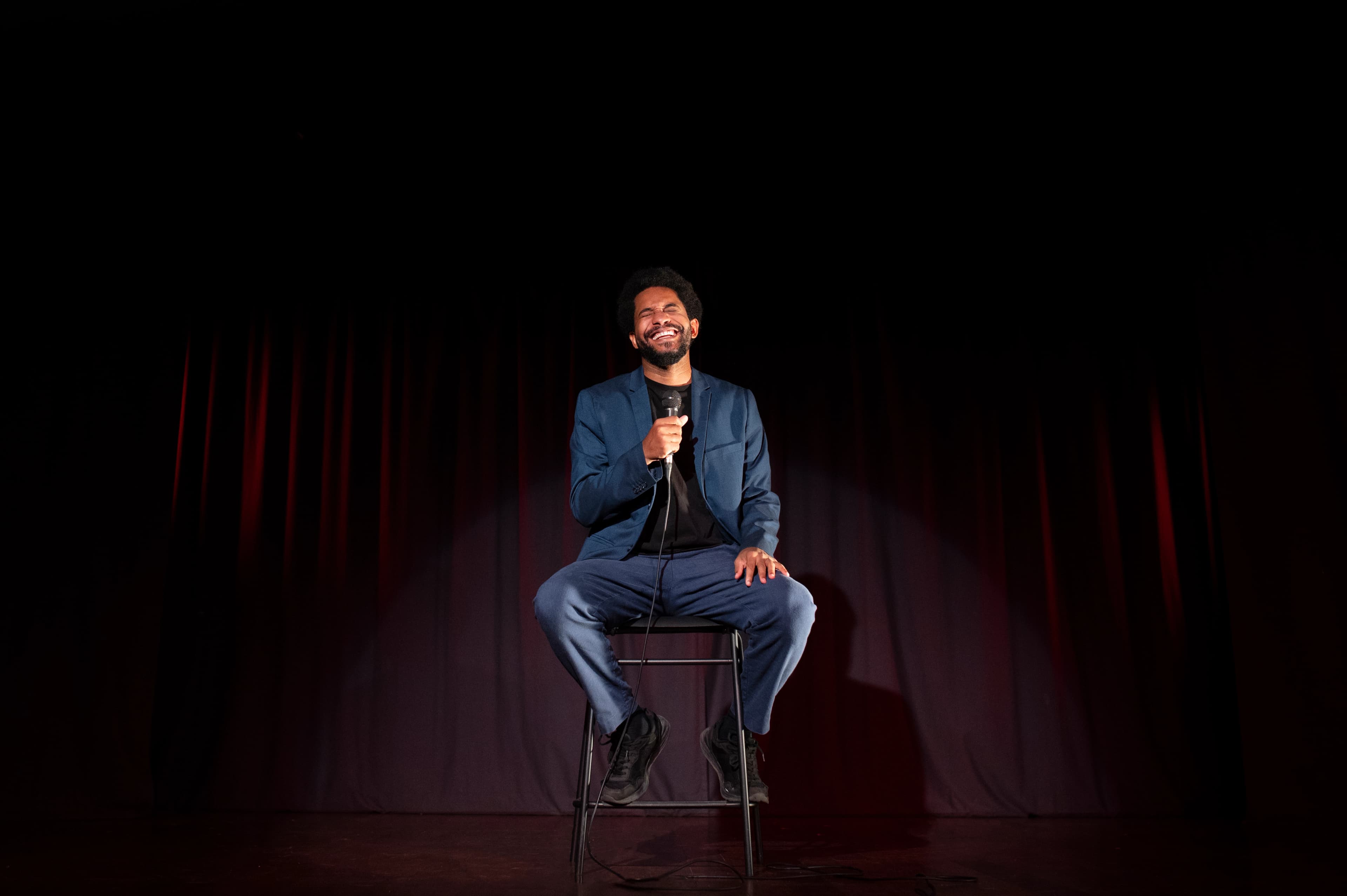 Stand-up comedian on stage with microphone, performing to an engaged audience in a comedy club setting.